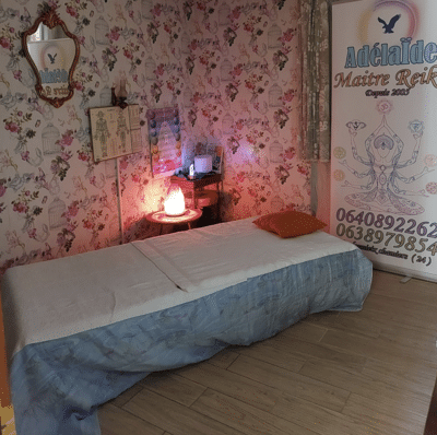Salle de soin Reiki avec table de massage, papier peint fleuri, lampe de sel rose, et bannière "Adélaïde Maître Reiki" avec numéros de téléphone.